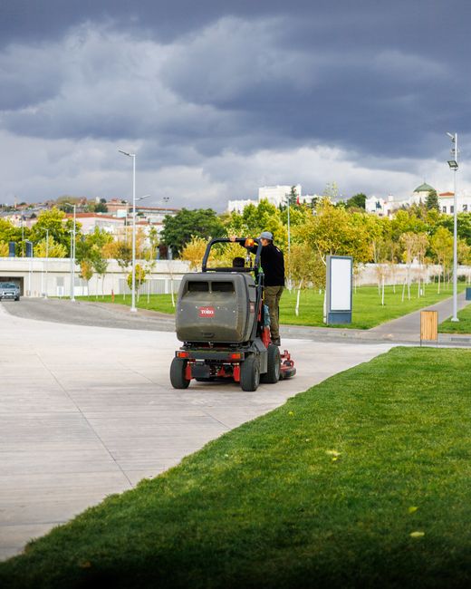 A person operating a ride-on lawn mower on a paved pathway in a well-maintained outdoor area, with trimmed grass in the foreground and a background featuring young trees, green lawns, and a parking lo