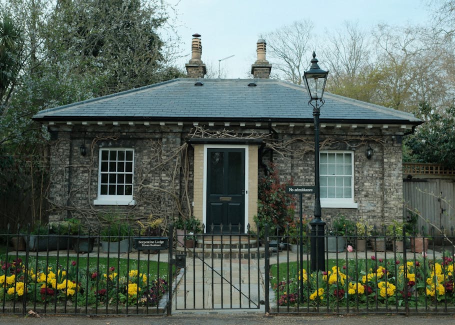 A small, single-story brick house with a dark slate roof, featuring two white-framed sash windows and a black front door accessed via a short brick staircase. The house is partially covered with leafl